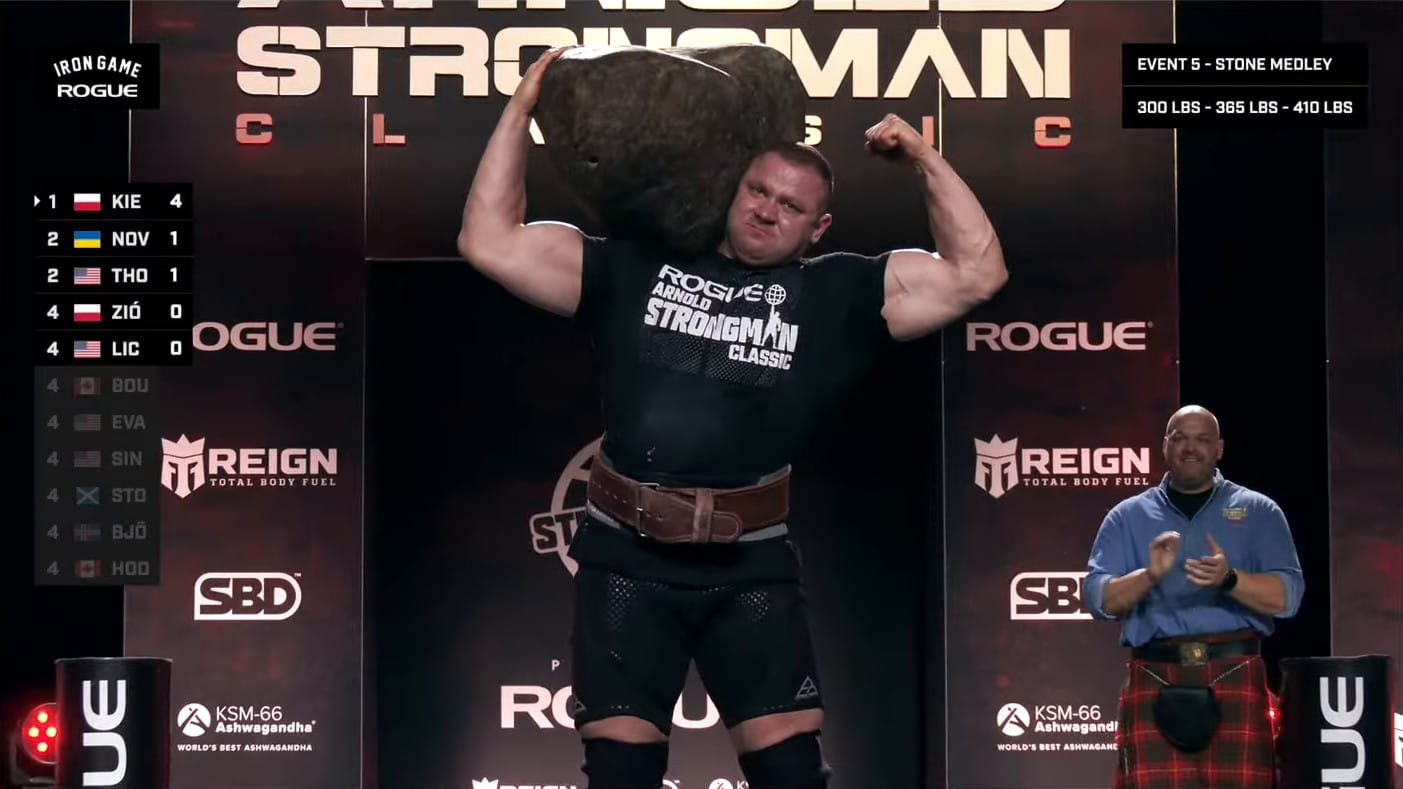 A screenshot from the Arnold Strongman Classic livestream. Mateusz Kieliszkowski stands with Odd Haugen's Tombstone on his shoulder. He flexes his other bicep.
