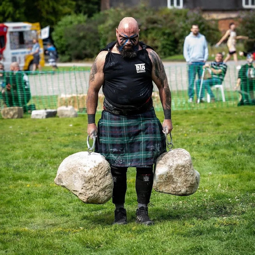 A man with facepaint carries a pair of ringed stones along a grass course.
