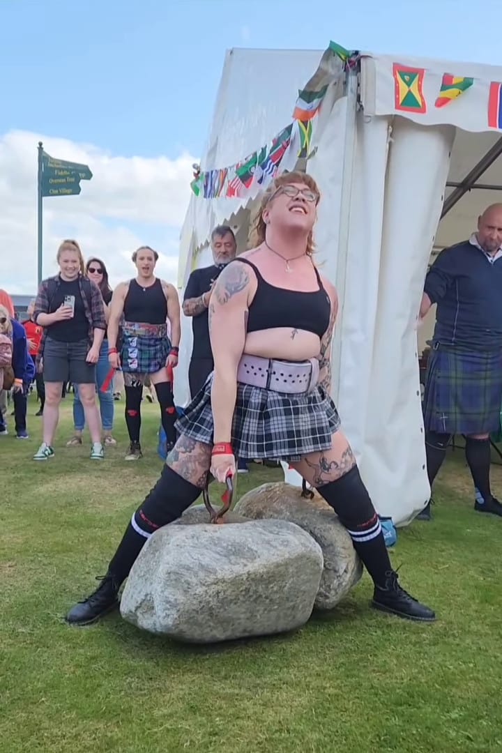 A woman stands with the Dinnie Stones between her legs, off the ground.