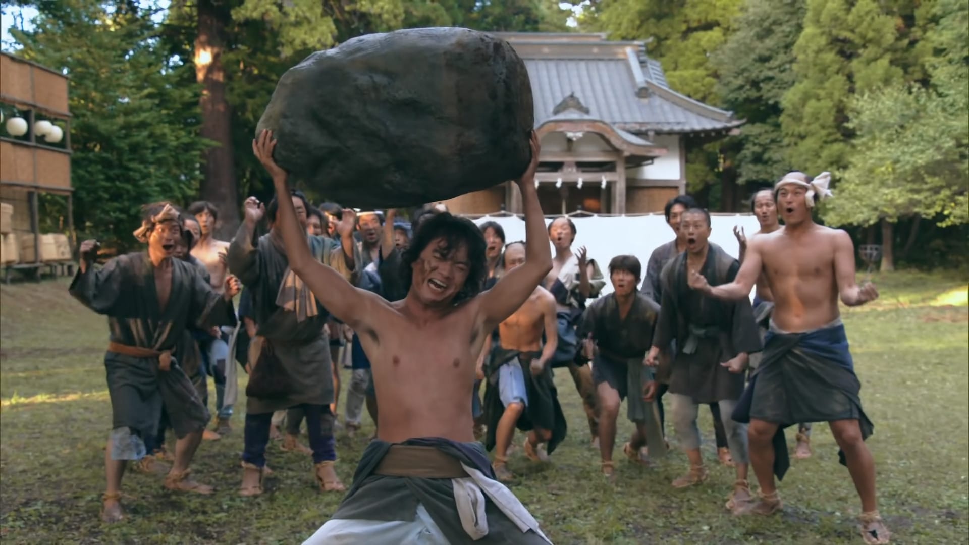 Ushi grimaces as he presses an enormous stone above his head. A crowd of peasants are cheering in the background.