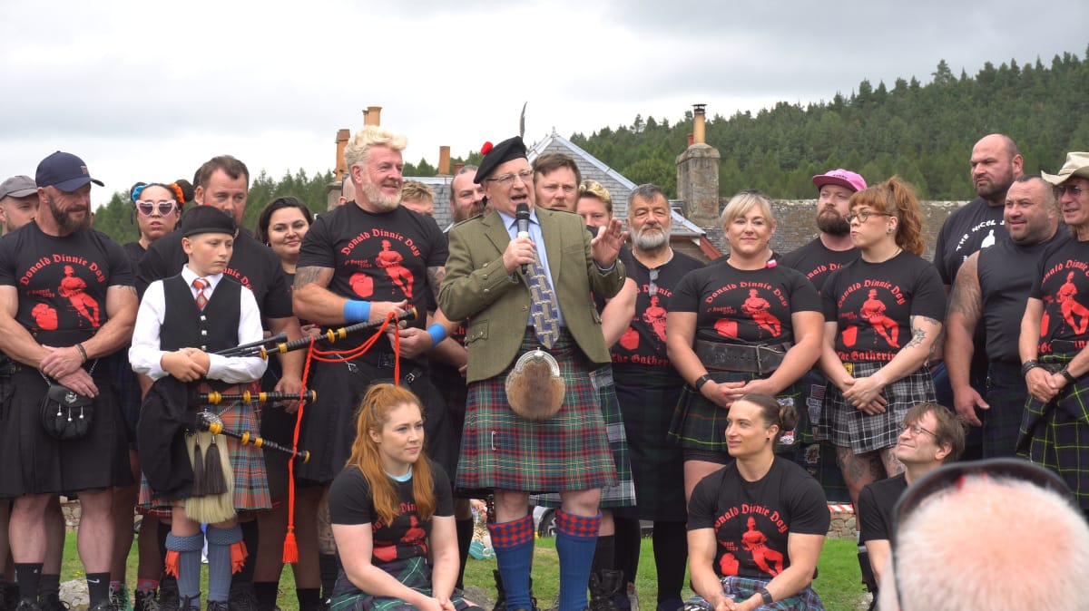 A kilted man wearing a hat topped with a feather gives an opening speech among a group of athletes.