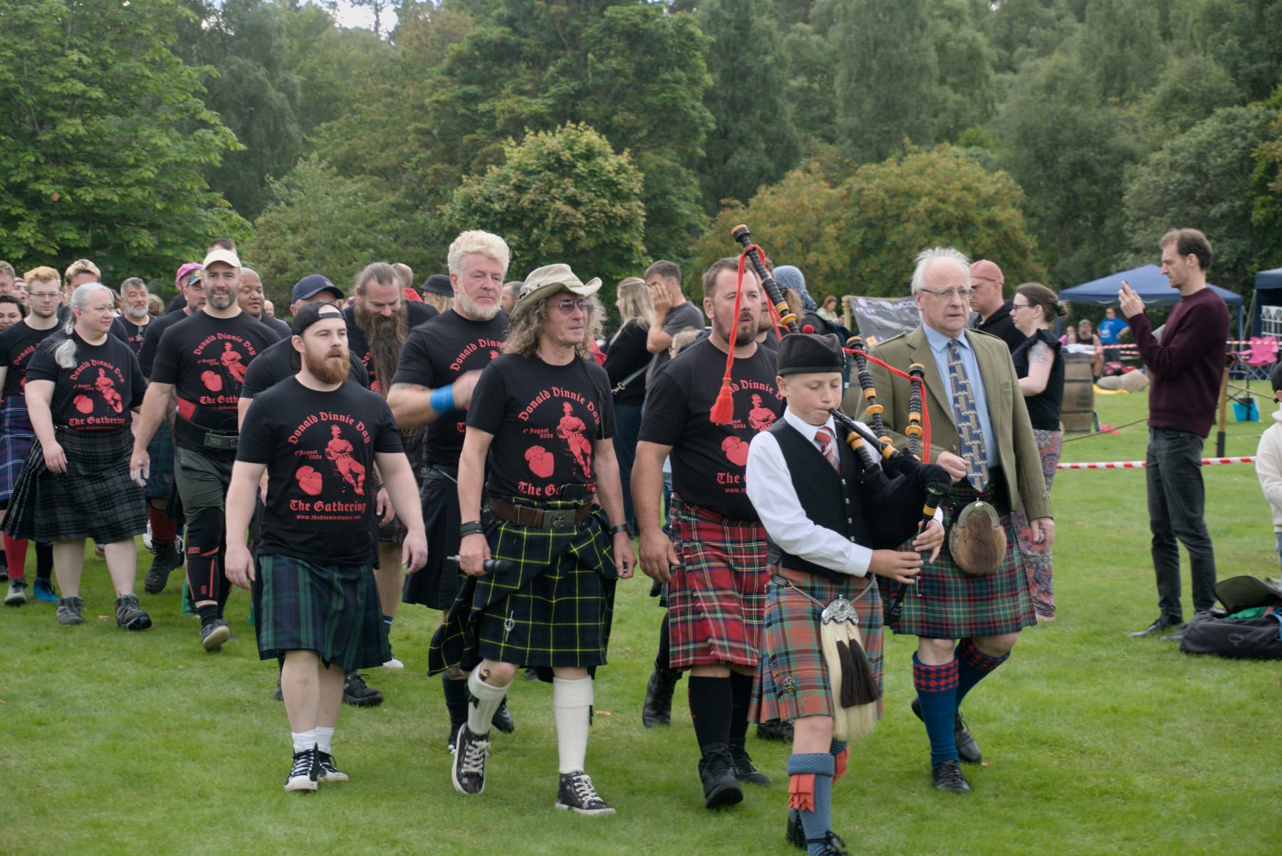 A large group of kilted athletes and officials are walking around Potarch green, lead by a young piper.