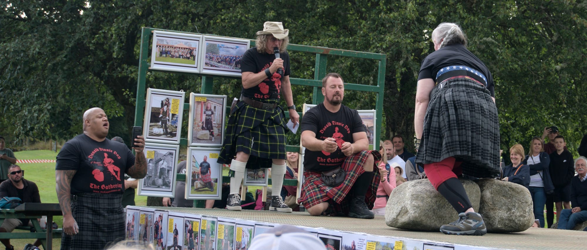 A woman attempts to lift the Dinnie Stones, her husband stands nearby, cheering and filming.