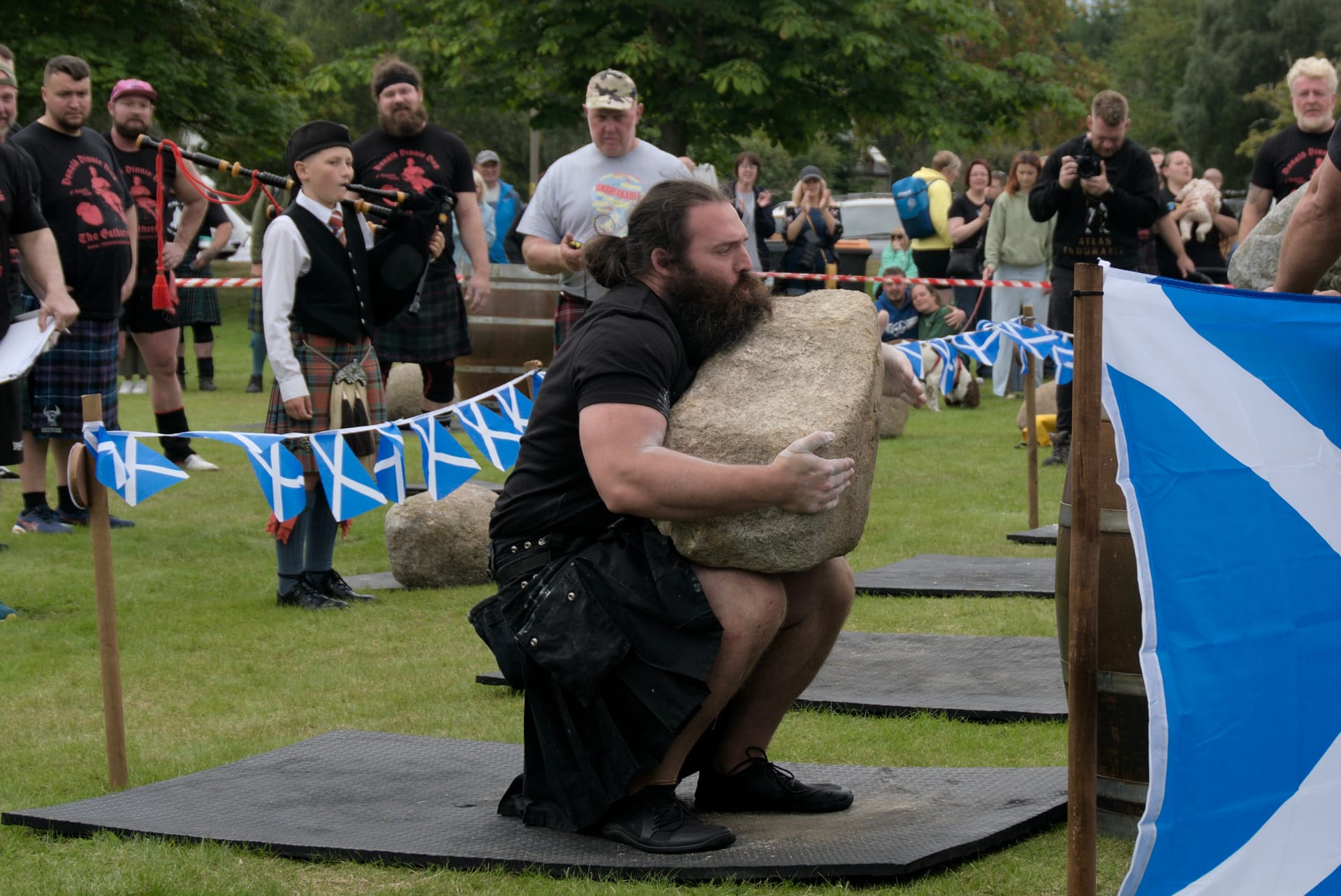 A man with long hair and a beard balances a large stone on his lap in front of a barrel.