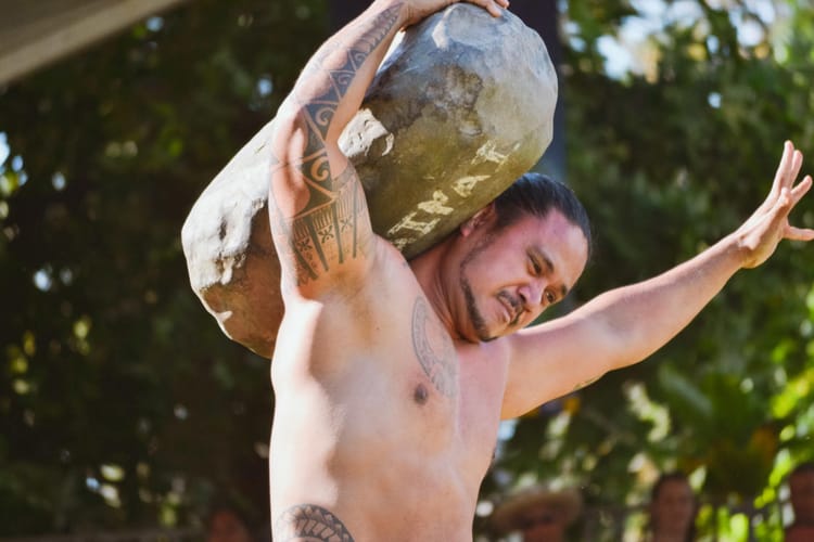 A man holds a long stone on his bare shoulder.